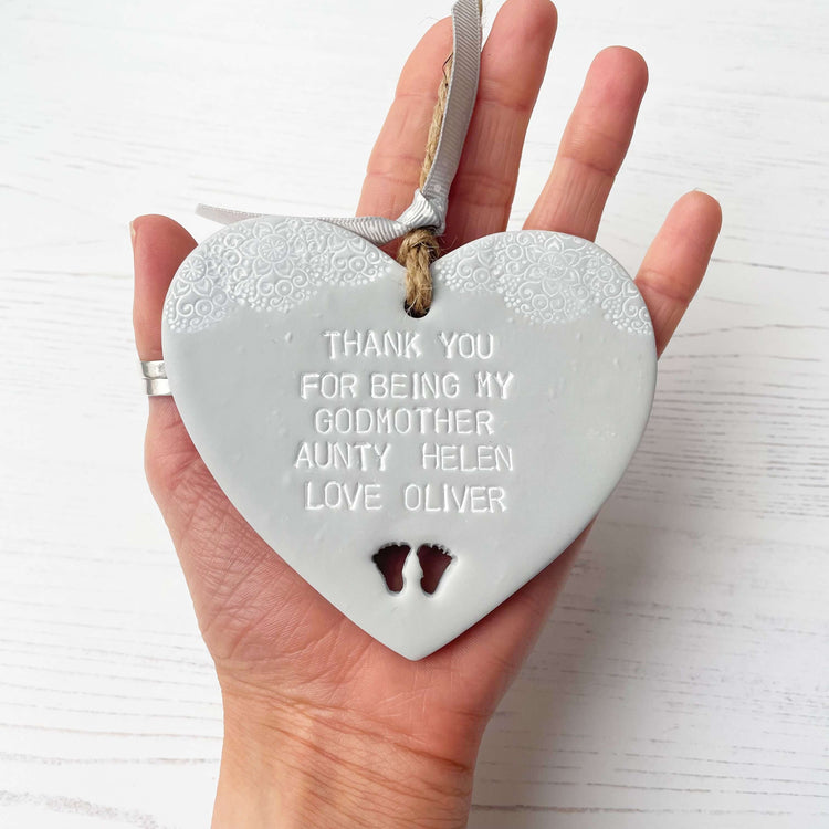 Personalised Godmother gift, grey clay hanging heart with baby feet cut out of the bottom and white lace edge at the top of the heart, the heart is personalised with THANK YOU FOR BEING MY GODMOTHER AUNTIE SARAH. LOVE WREN 23/06/2024