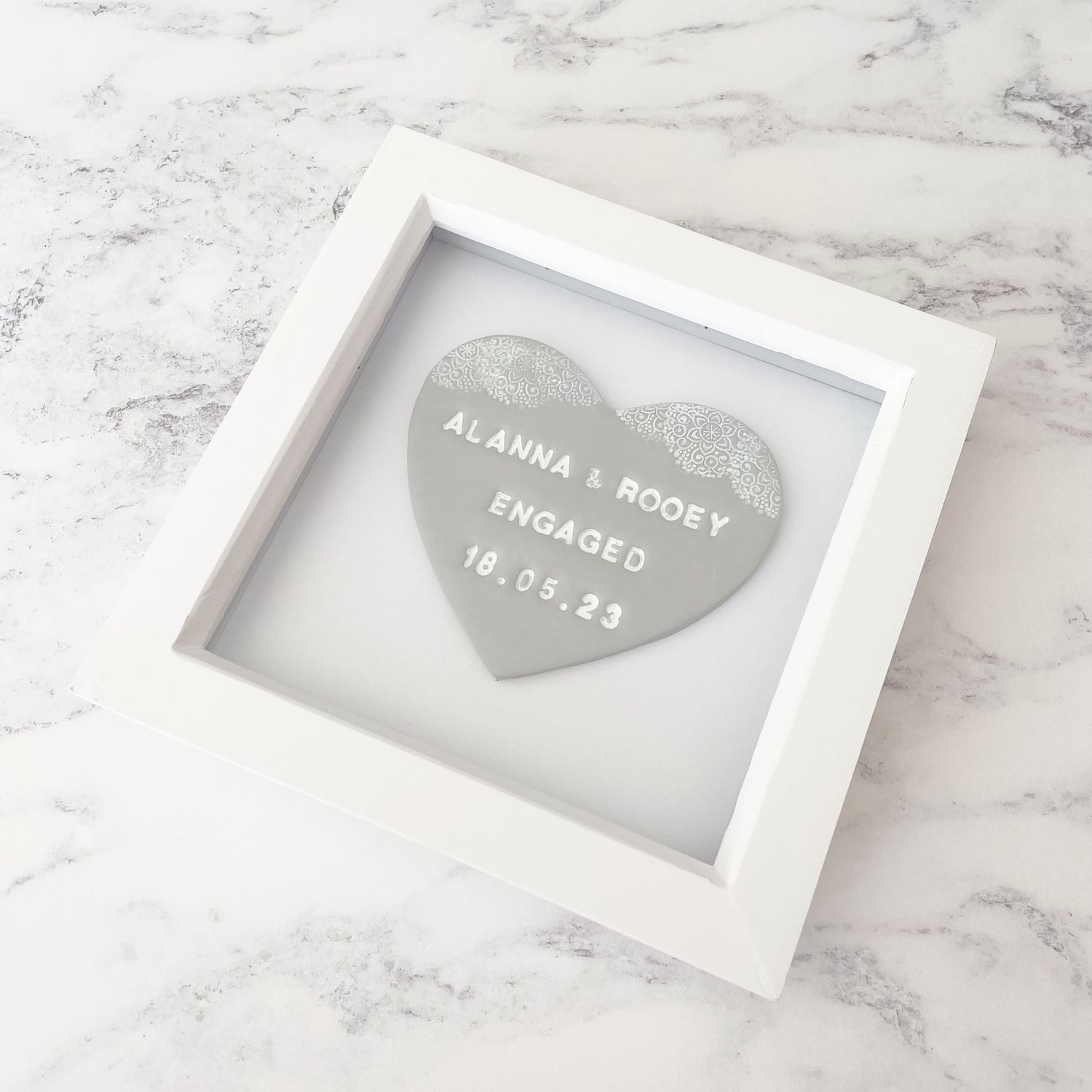 Personalised framed engagement gift, grey clay heart with a white lace edge at the top of the heart in a white box frame, the heart is personalised with ALANNA & ROOEY ENGAGED 18.05.23