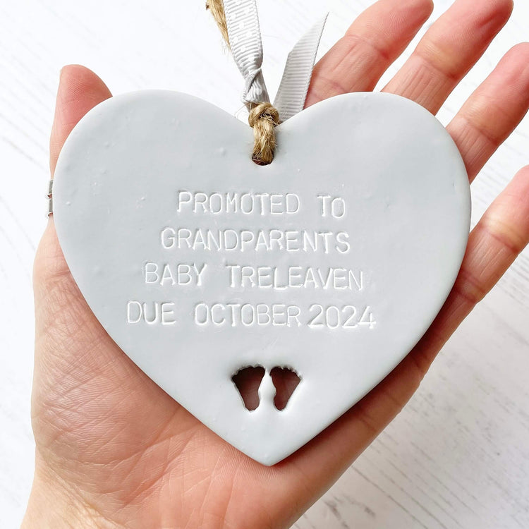 Grey clay hanging heart with baby feet cut out of the bottom and grey personalisation, the heart is personalised with PROMOTED TO GRANDPARENTS BABY TRELEAVEN DUE OCTOBER 2024