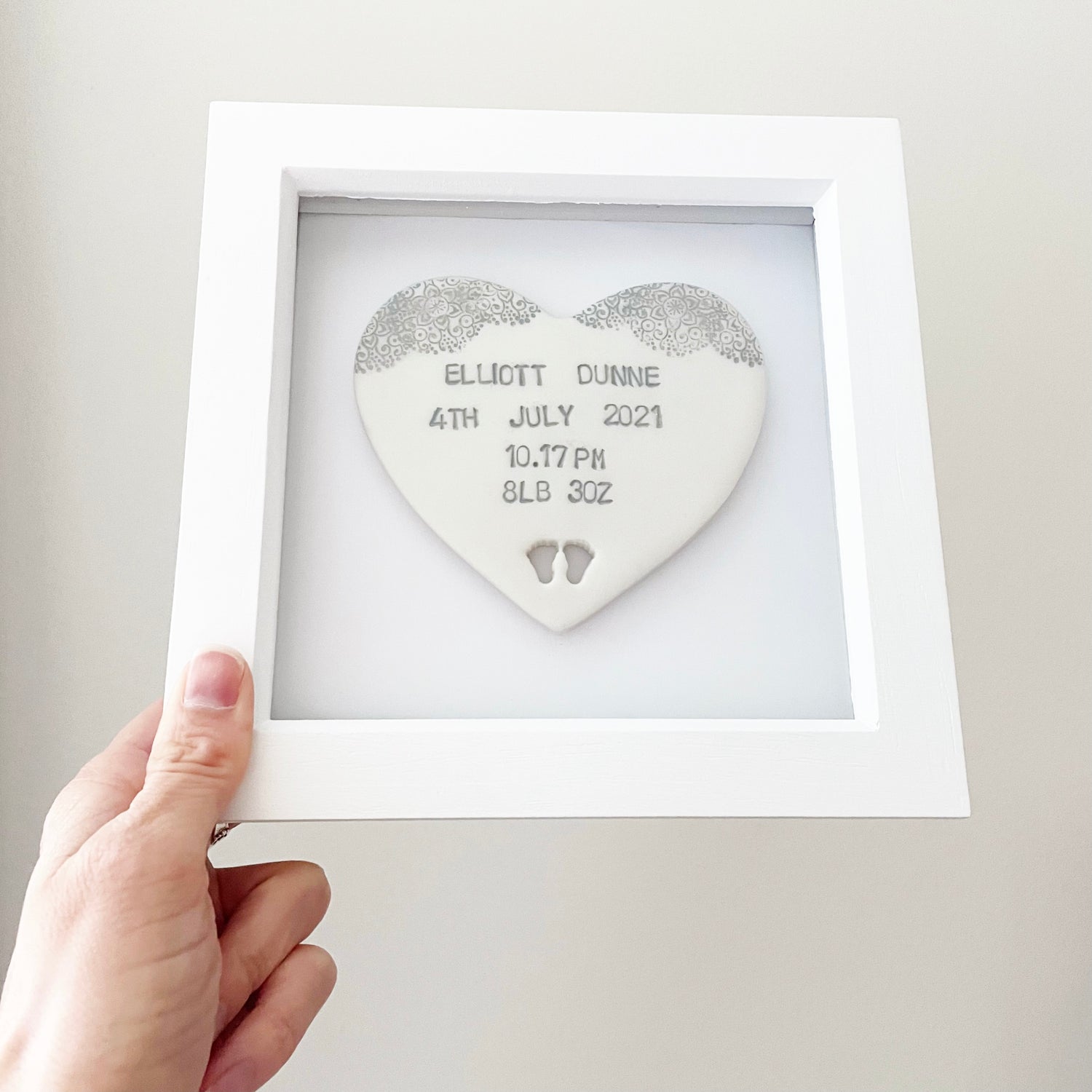 Personalised baby keepsake gift, pearlised white clay heart with baby feet cut out at the bottom of the heart and a grey lace edge at the top of the heart in a white box frame, the heart is personalised with the baby’s name, date of birth, weight and time