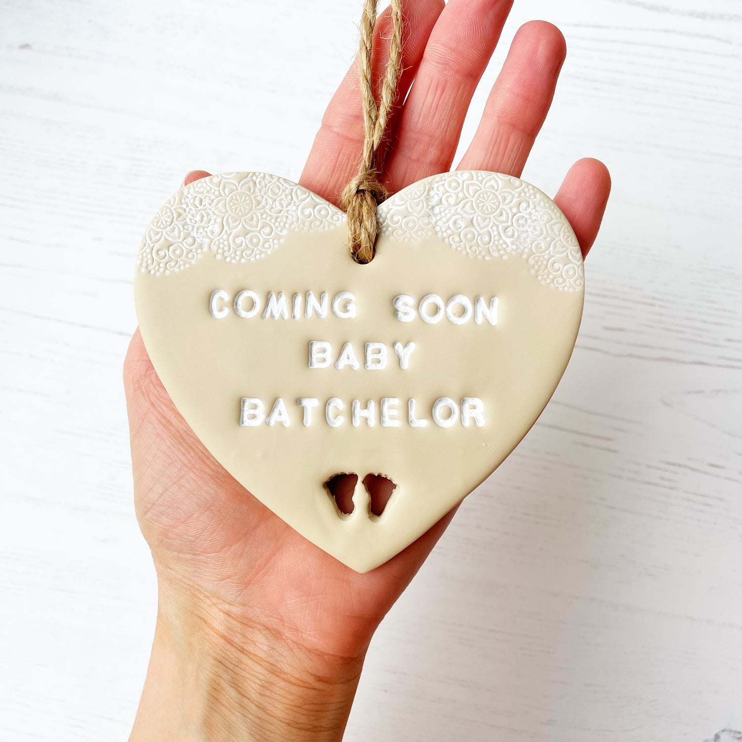Personalised pregnancy reveal sign keepsake, beige clay hanging heart with a white lace edge at the top of the heart and baby feet cut out at the bottom, the heart is personalised with COMING SOON BABY BATCHELOR