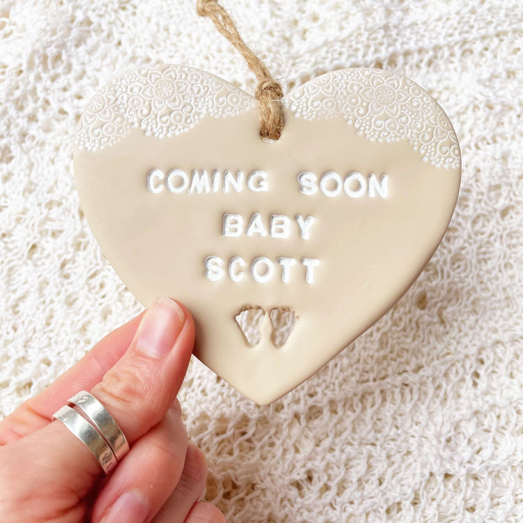 Personalised pregnancy reveal sign keepsake, beige clay hanging heart with a white lace edge at the top of the heart and baby feet cut out at the bottom, the heart is personalised with COMING SOON BABY BATCHELOR