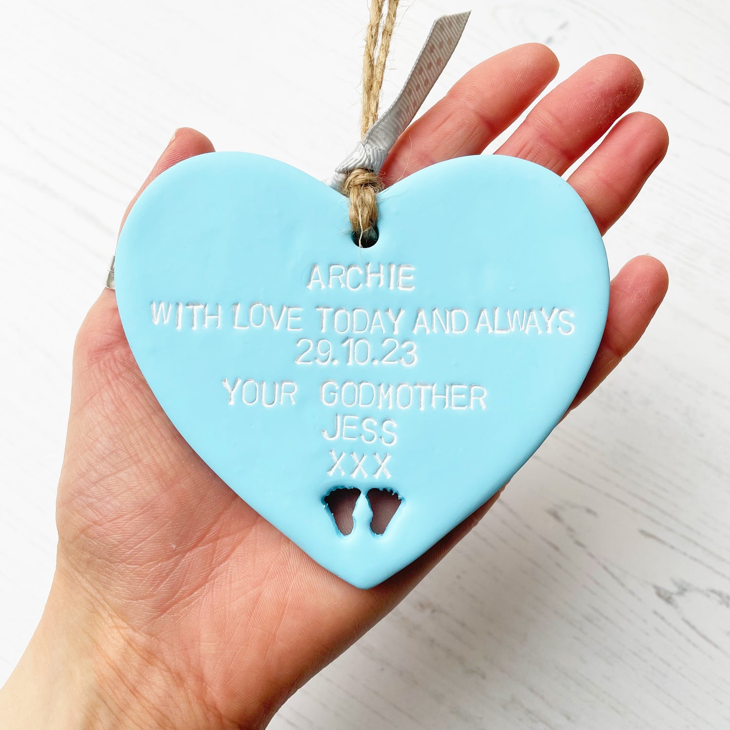 Personalised Christening gift, pastel blue clay hanging heart with baby feet cut out of the bottom, the heart is personalised with ARCHIE WITH LOVE TODAY AND ALWAYS 29.10.23 YOUR GODMOTHER JESS XXX