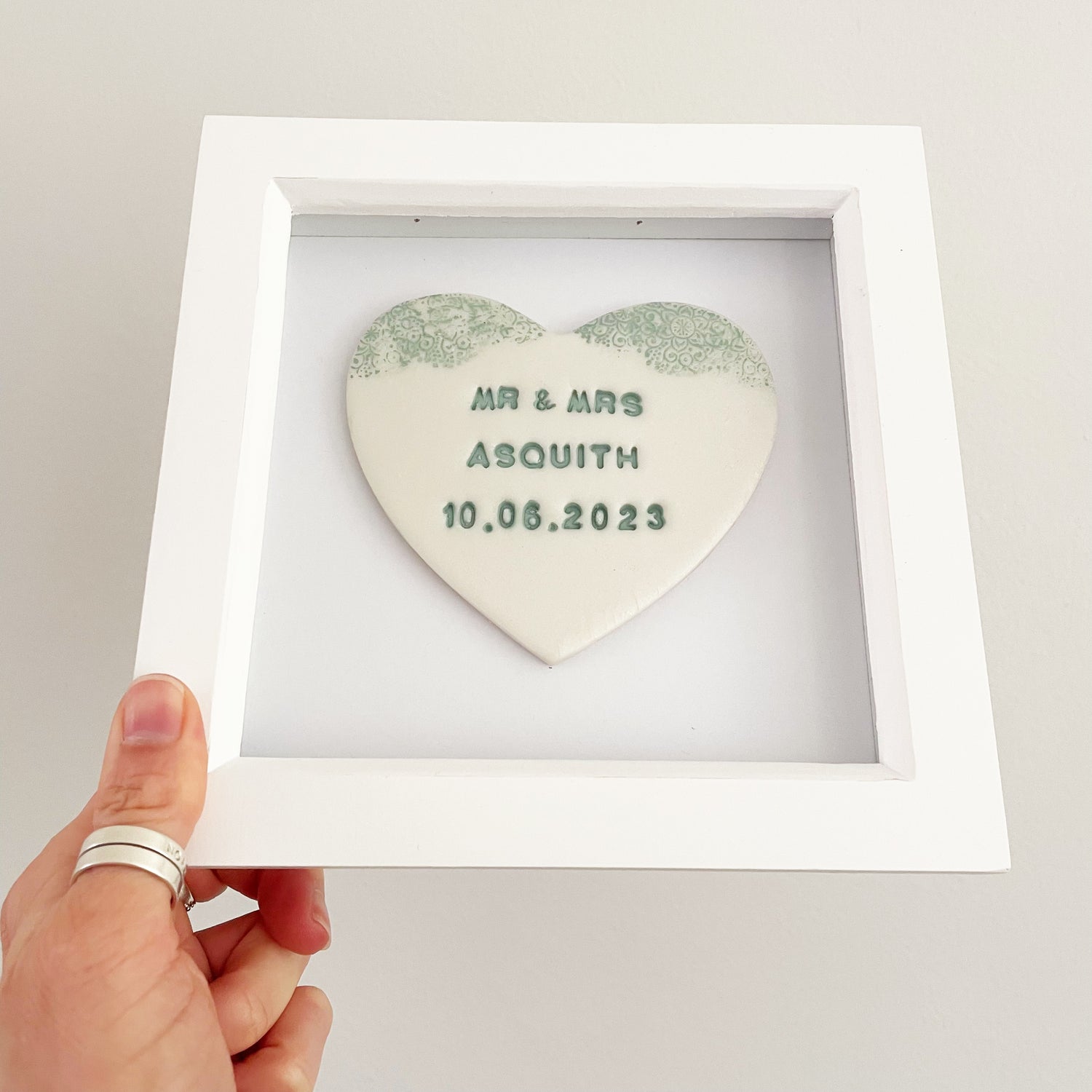 Personalised hanging wedding heart gift, pearlised white clay heart with a sage green lace edge at the top of the heart in a white box frame, the heart is personalised with MR & MRS ASQUITH 10.06.2023