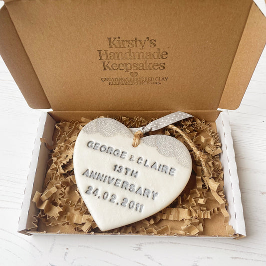 Personalised 13th anniversary gift, pearlised white clay hanging heart with a grey lace edge at the top of the heart, the heart is personalised with GEORGE & CLAIRE 13TH ANNIVERSARY 24.02.2011 in a postal box with brown kraft shredded zigzag paper