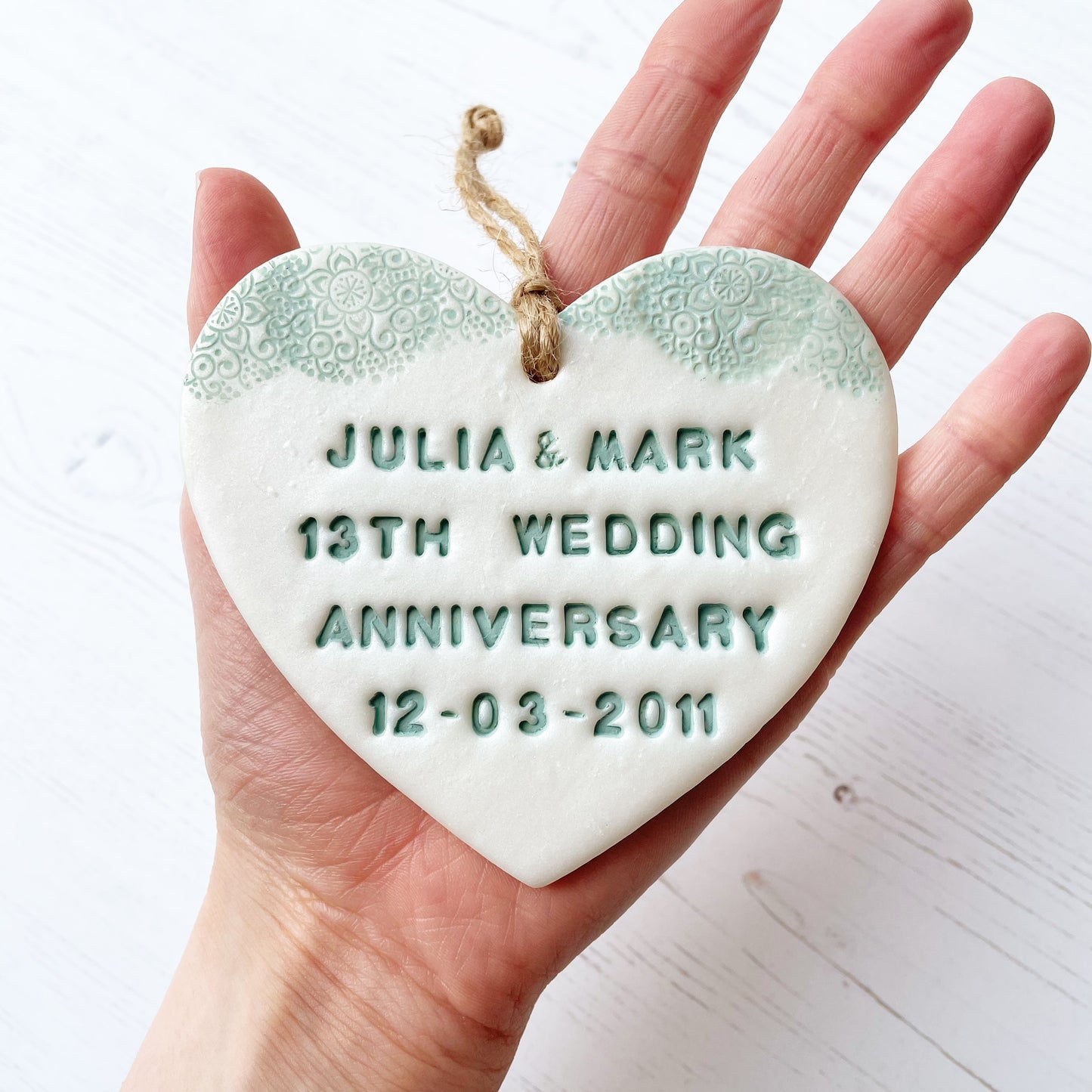 Personalised 13th wedding anniversary gift, pearlised white clay hanging heart with a sage green lace edge at the top of the heart, the heart is personalised with JULIA & MARK 13TH WEDDING ANNIVERSARY 12.03.2011