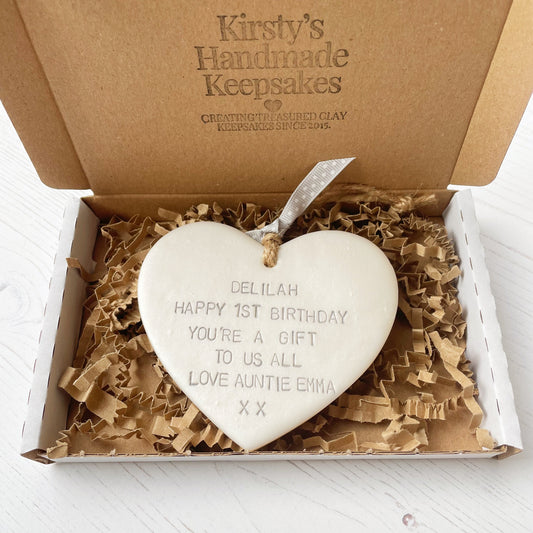 Personalised 1st birthday gift, pearlised white clay hanging heart, the heart is personalised in grey with DELILAH HAPPY 1ST BIRTHDAY YOU’RE A GIFT TO US ALL LOVE AUNTIE EMMA XX in a postal box with brown Kraft shredded zigzag paper