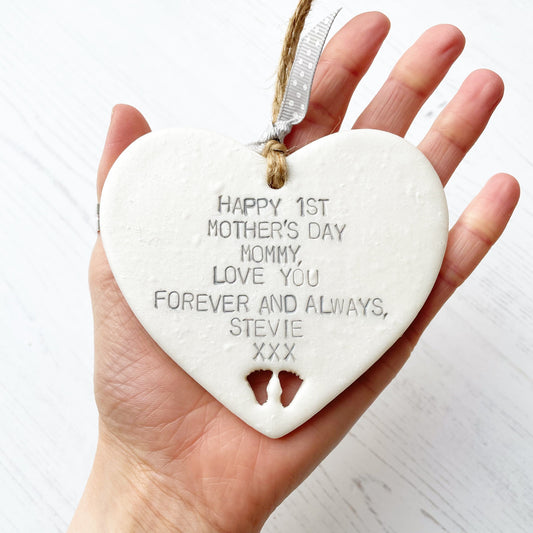 Personalised first Mother’s Day gift, pearlised white hanging clay heart with baby feet cut out at the bottom of the heart, the heart is personalised in grey with HAPPY 1ST MOTHER'S DAY MOMMY, LOVE YOU FOREVER AND ALWAYS, STEVIE XXX