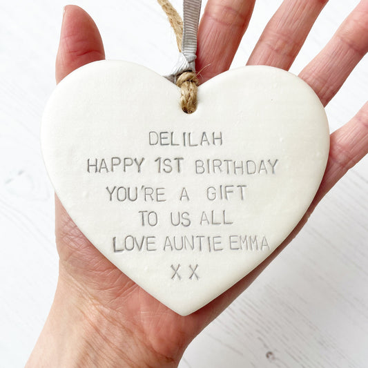 Personalised 1st birthday gift, pearlised white clay hanging heart, the heart is personalised in grey with DELILAH HAPPY 1ST BIRTHDAY YOU’RE A GIFT TO US ALL LOVE AUNTIE EMMA XX