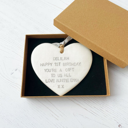 Personalised 1st birthday gift, pearlised white clay hanging heart, the heart is personalised in grey with DELILAH HAPPY 1ST BIRTHDAY YOU’RE A GIFT TO US ALL LOVE AUNTIE EMMA XX in a brown Kraft luxury gift box