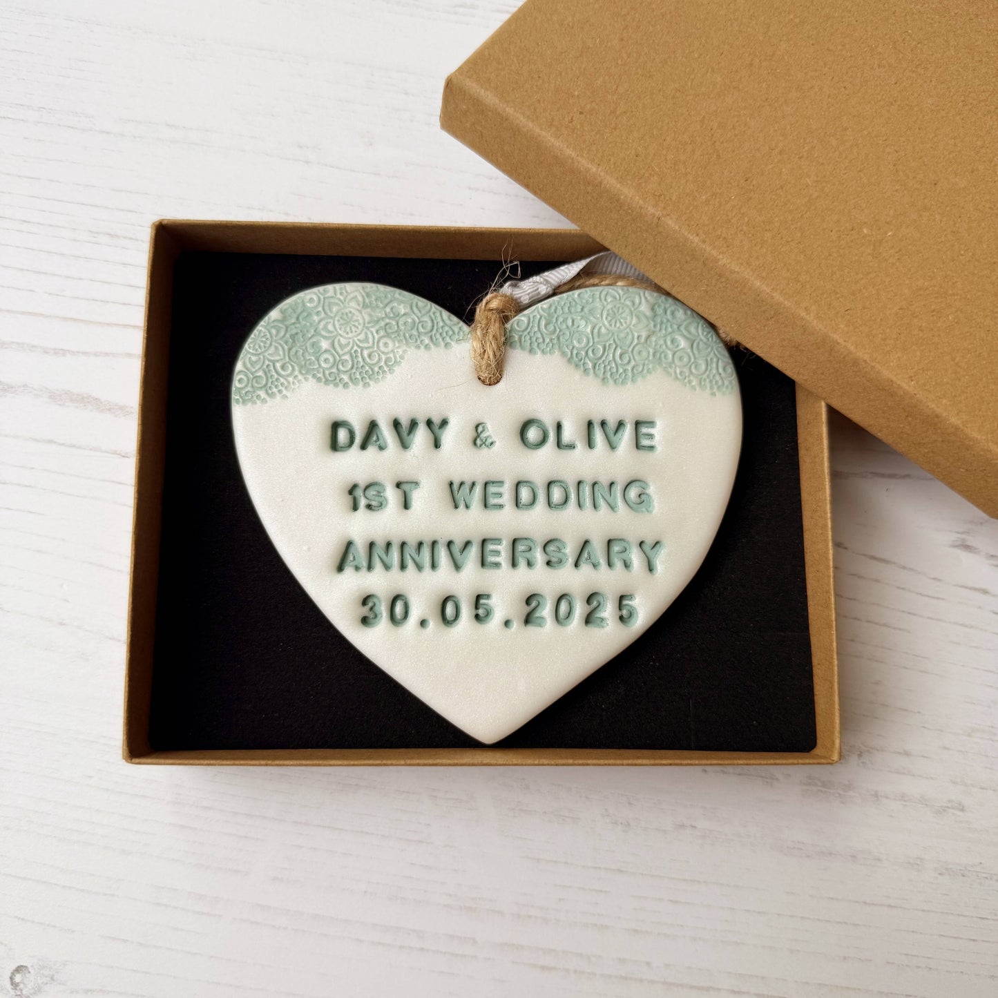 Personalised 1st wedding anniversary gift, pearlised white clay hanging heart with a sage green lace edge at the top of the heart, the heart is personalised with DAVY & OLIVE 1ST WEDDING ANNIVERSARY 30.05.2025