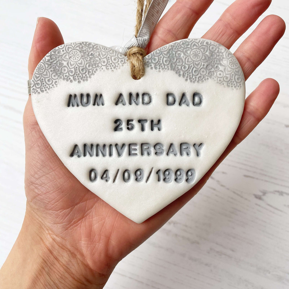 Personalised 25th anniversary gift, pearlised white clay hanging heart with a grey lace edge at the top of the heart, the heart is personalised with MUM AND DAD 25TH ANNIVERSARY 04/09/1999