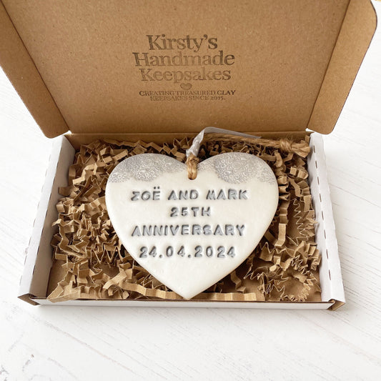 Personalised 25th anniversary gift, pearlised white clay hanging heart with a grey lace edge at the top of the heart, the heart is personalised with ZOË AND MARK 25TH ANNIVERSARY 24.04.2024 In a postal box with brown Kraft shredded zigzag paper