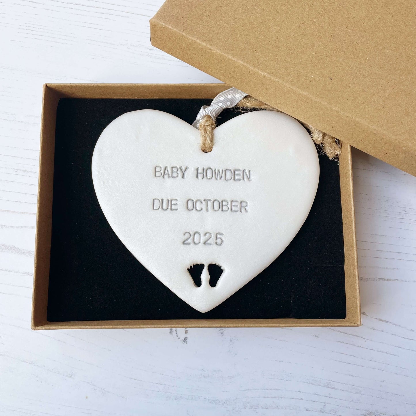 Pearlised white clay hanging heart with baby feet cut out of the bottom and grey personalisation, the heart is personalised with BABY HOWDEN DUE OCTOBER 2025