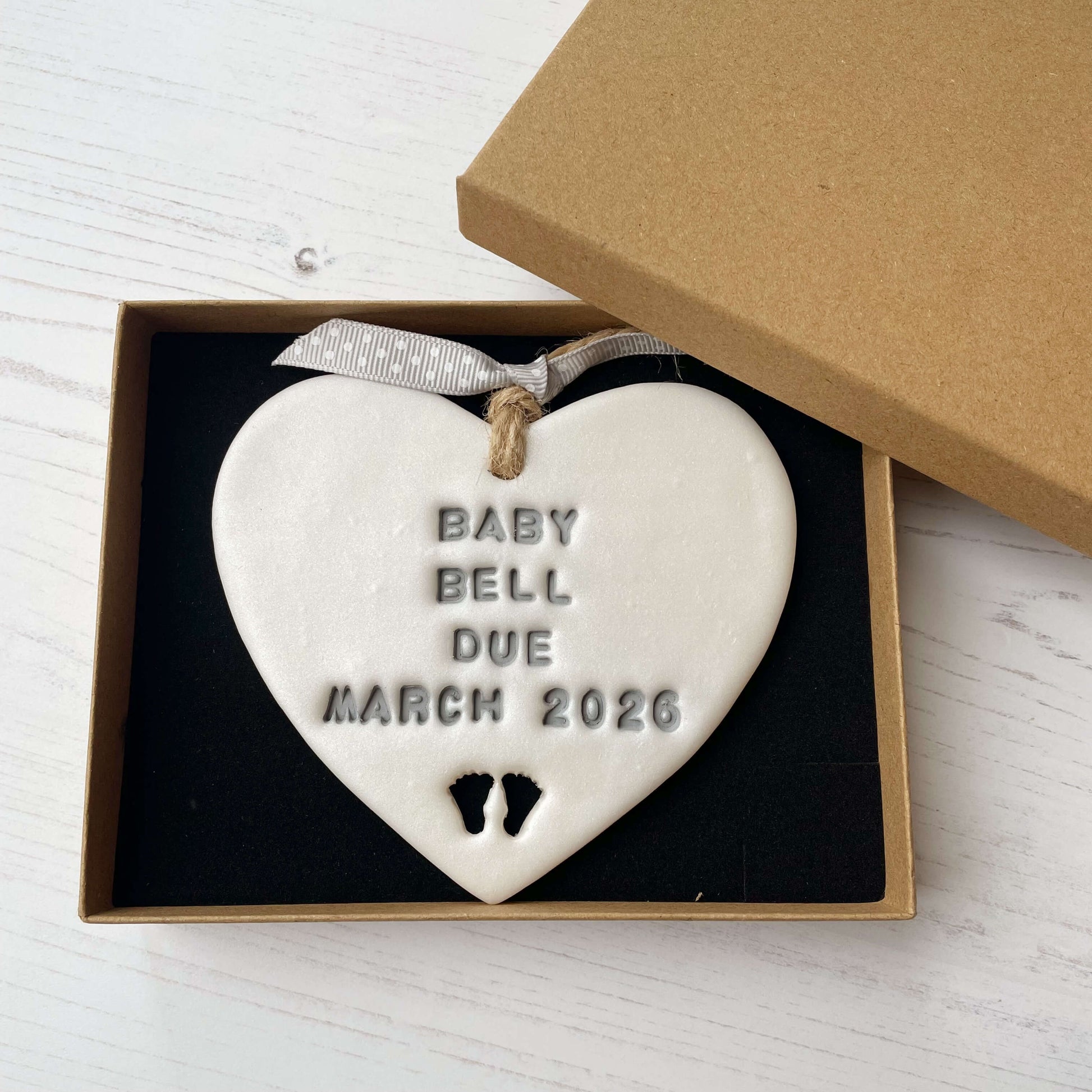 Personalised pregnancy reveal sign keepsake, pearlised white clay hanging heart with baby feet cut out at the bottom, the heart is personalised in grey with BABY BELL DUE MARCH 2026