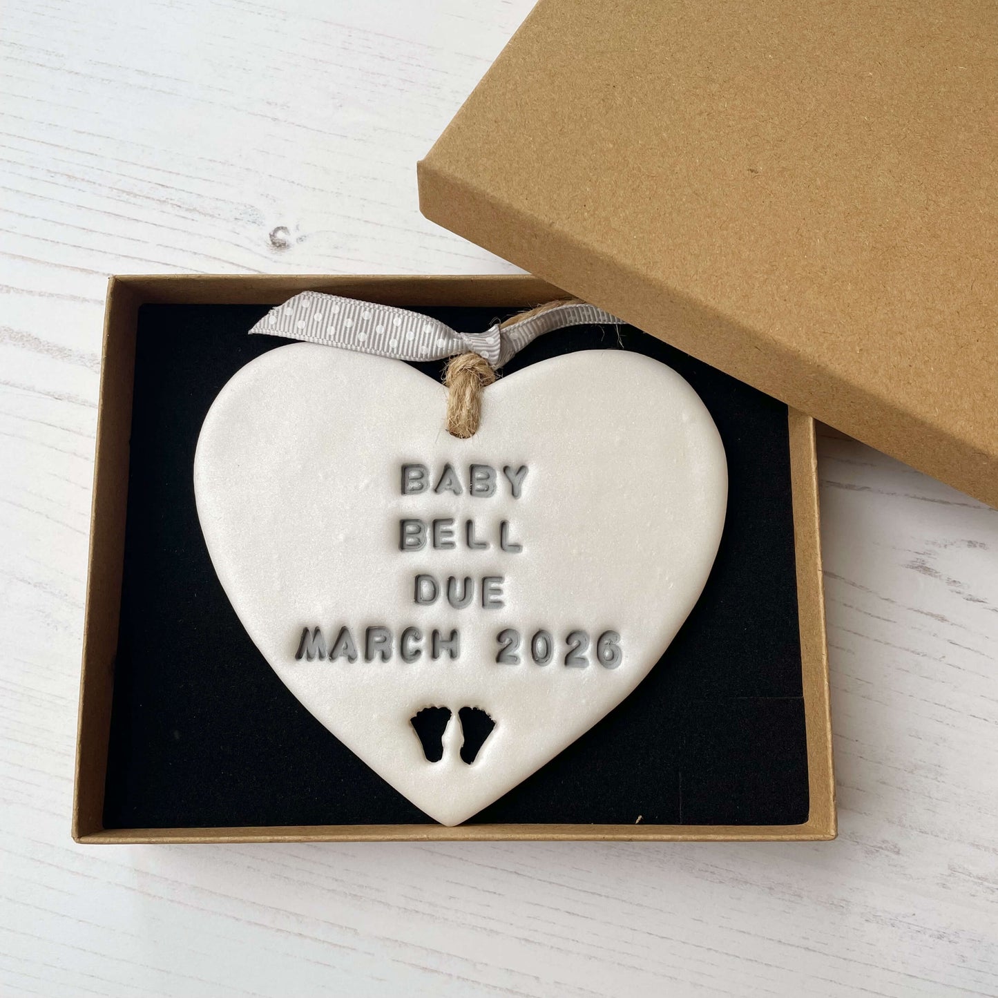 Personalised pregnancy reveal sign keepsake, pearlised white clay hanging heart with baby feet cut out at the bottom, the heart is personalised in grey with BABY BELL DUE MARCH 2026