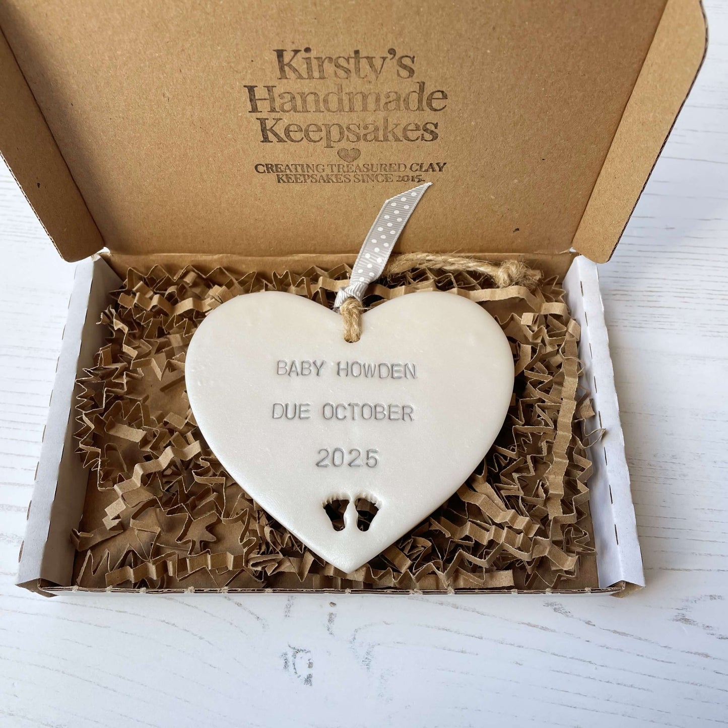 Pearlised white clay hanging heart with baby feet cut out of the bottom and grey personalisation, the heart is personalised with BABY HOWDEN DUE OCTOBER 2025