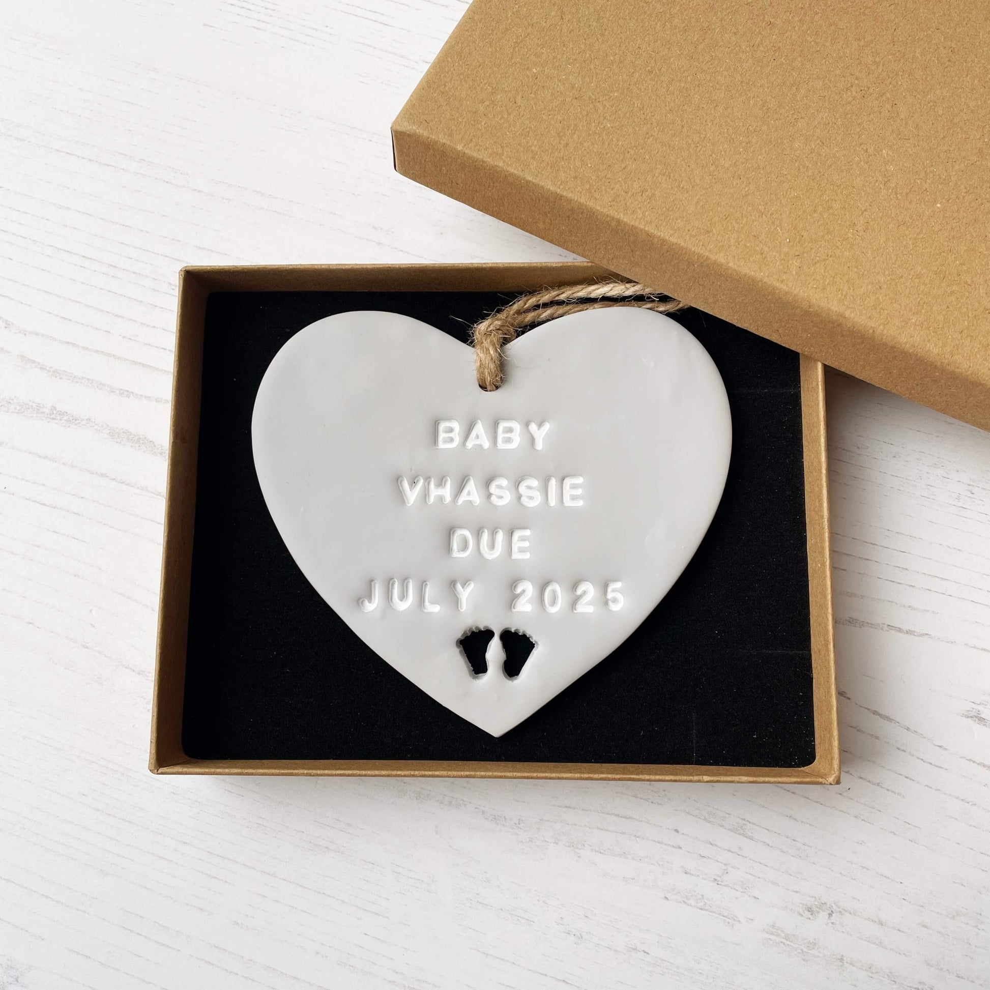 Personalised pregnancy reveal sign keepsake, grey clay hanging heart with baby feet cut out at the bottom, the heart is personalised in white with BABY VHASSIE DUE JULY 2025 In a white postal box with brown Kraft shredded zigzag paper