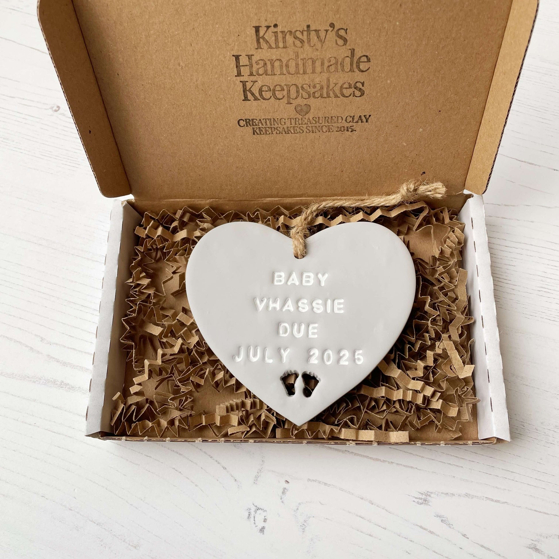 Personalised pregnancy reveal sign keepsake, grey clay hanging heart with baby feet cut out at the bottom, the heart is personalised in white with BABY VHASSIE DUE JULY 2025 In a white postal box with brown Kraft shredded zigzag paper