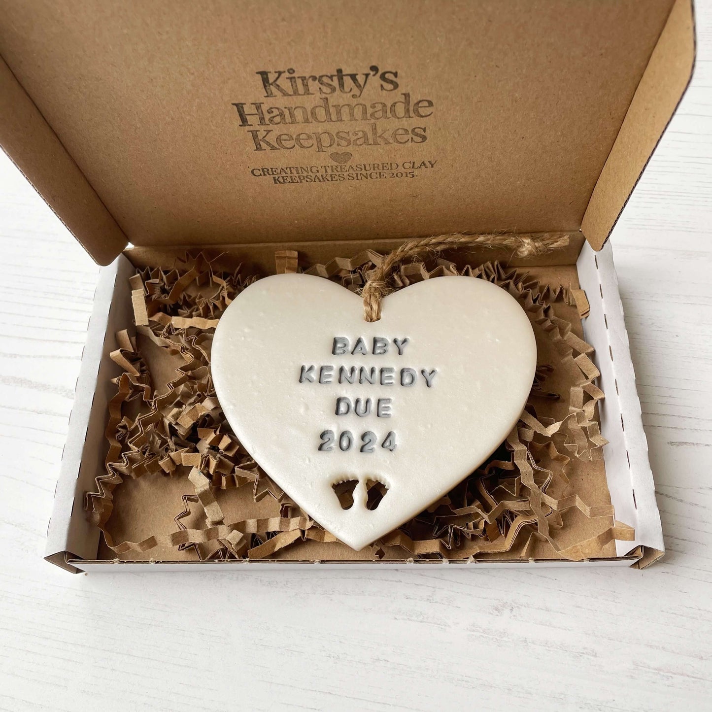 Personalised pregnancy reveal sign keepsake, pearlised white clay hanging heart with baby feet cut out at the bottom, the heart is personalised in grey with BABY KENNEDY DUE 2024 In a white postal box with brown Kraft shredded zigzag paper