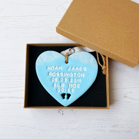 Personalised baby gift pastel blue clay hanging heart with a white lace edge top baby feet cut out at the bottom the heart is personalised with the baby’s name, date of birth, weight and time In a brown Kraft luxury gift box
