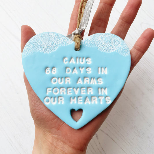 Personalised baby memorial keepsake gift, blue clay heart with a white lace edge at the top of the heart and a heart cut out at the bottom with jute twine for hanging, the heart is personalised with CAIUS 68 DAYS IN OUR ARMS FOREVER IN OUR HEARTS
