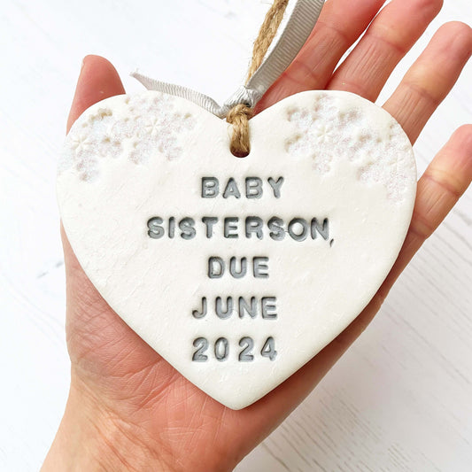 Personalised baby's first Christmas heart ornament, pearlised white clay with BABY SISTERSON, DUE JUNE 2024 painted grey, decorated with 2 iridescent glitter snowflakes on either side of the top of the heart