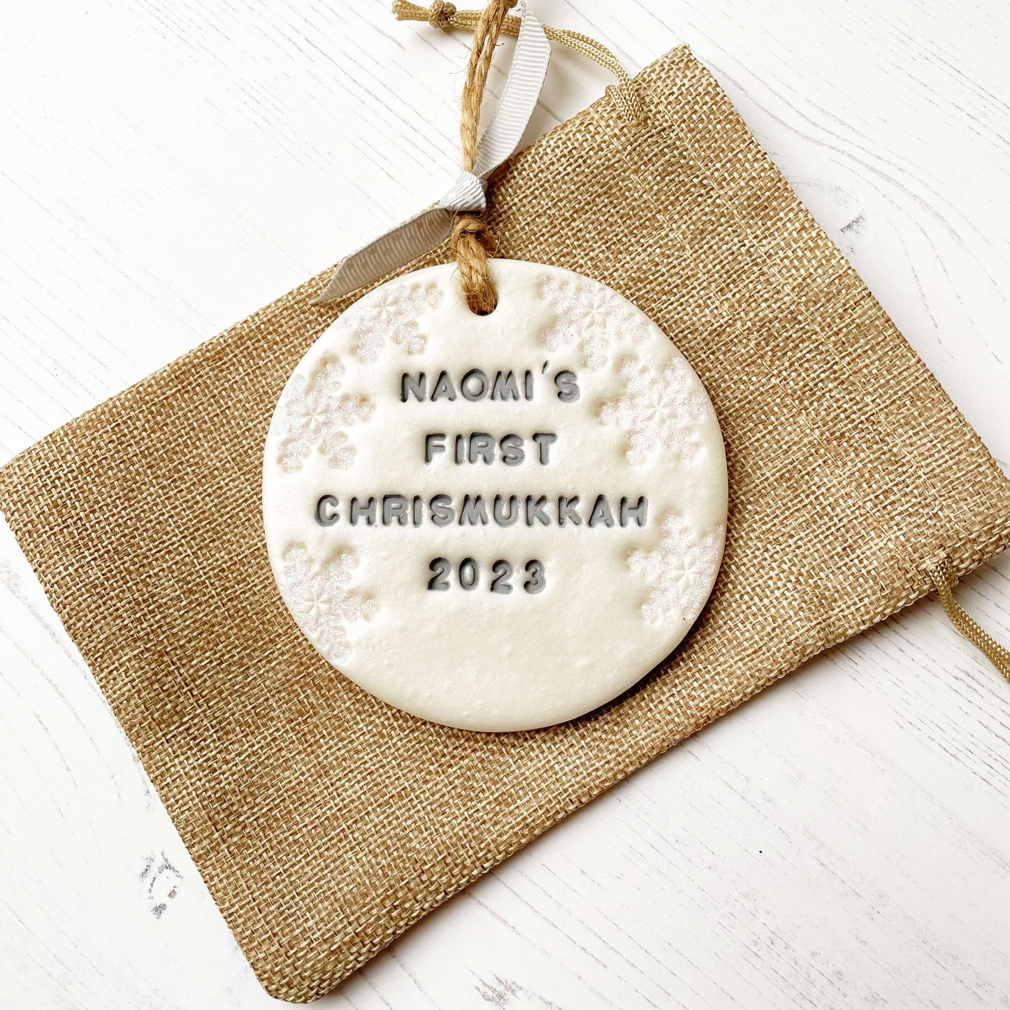 Personalised baby’s first Christmas bauble ornament, pearlised white clay round personalised with NAOMI’S FIRST CHRISMUKKAH 2023 in grey paint, decorated with 3 iridescent glitter snowflakes on either side of the bauble