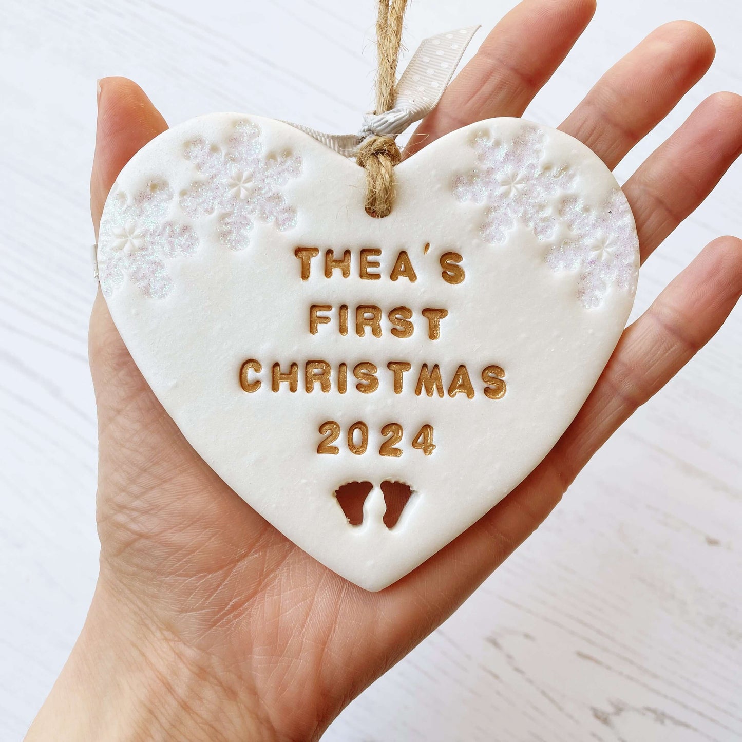 Personalised baby’s first Christmas heart ornament, pearlised white clay with THEA’S FIRST CHRISTMAS 2024 painted gold, decorated with 2 iridescent glitter snowflakes on either side of the top of the heart and baby feet cut out at the bottom