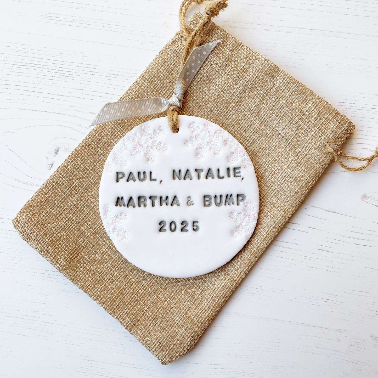 Personalised bump’s first Christmas bauble ornament, glitter white round clay with PAUL, NATALIE, MARTHA & BUMP 2025 in grey paint, decorated with 3 iridescent glitter snowflakes on either side of the bauble