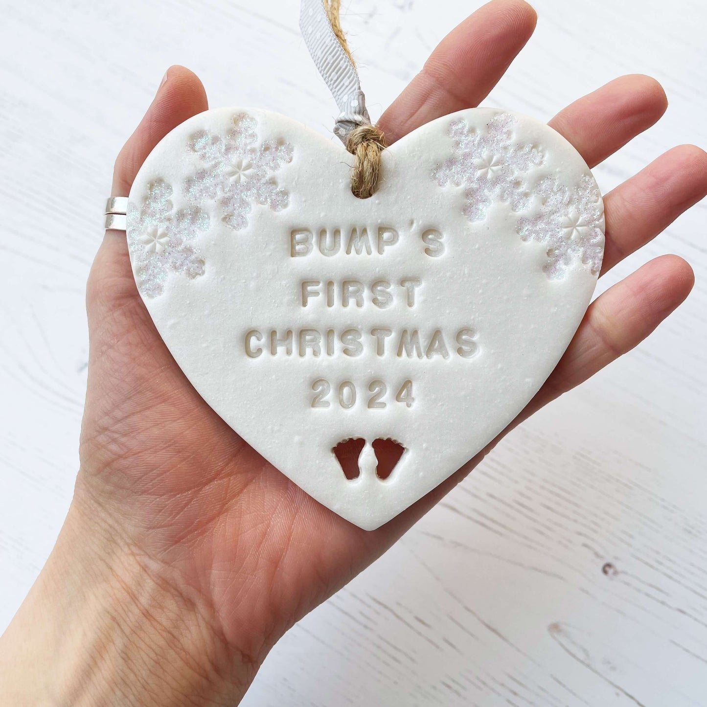 Personalised baby’s first Christmas heart ornament, pearlised white clay with ALVA’S FIRST CHRISTMAS 2024, decorated with 2 iridescent glitter snowflakes on either side of the top of the heart and baby feet cut out at the bottom