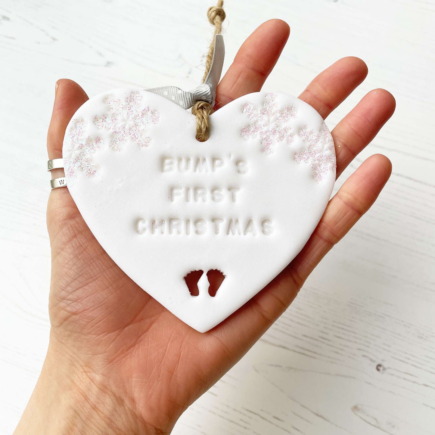 Personalised bump’s first Christmas heart ornament, glitter white clay with BUMP’S FIRST CHRISTMAS, decorated with 2 iridescent glitter snowflakes on either side of the top of the heart and baby feet cut out at the bottom