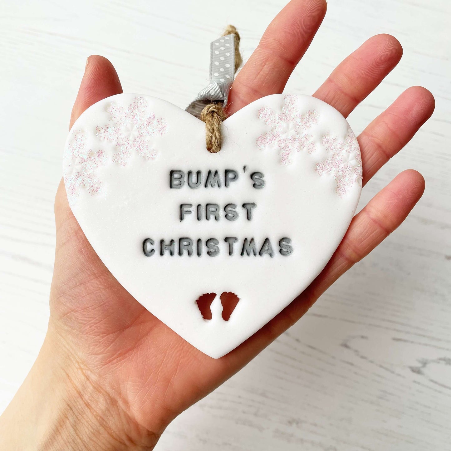 Personalised bump’s first Christmas heart ornament, glitter white clay with BUMP’S FIRST CHRISTMAS painted grey, decorated with 2 iridescent glitter snowflakes on either side of the top of the heart and baby feet cut out at the bottom