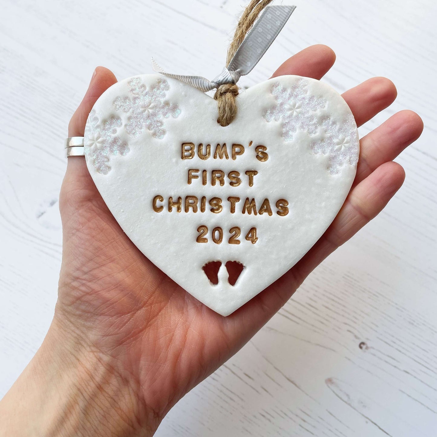 Personalised baby’s first Christmas heart ornament, pearlised white clay with THEA’S FIRST CHRISTMAS 2024 painted gold, decorated with 2 iridescent glitter snowflakes on either side of the top of the heart and baby feet cut out at the bottom