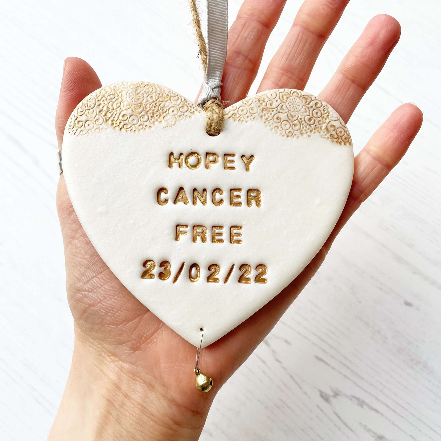 Personalised cancer survivor gift, pearlised white clay hanging heart with a gold lace edge at the top of the heart and a gold bell hanging below, the heart is personalised with HOPEY CANCER FREE 23/02/22