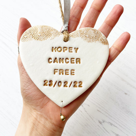 Personalised cancer survivor gift, pearlised white clay hanging heart with a gold lace edge at the top of the heart and a gold bell hanging below, the heart is personalised with HOPEY CANCER FREE 23/02/22