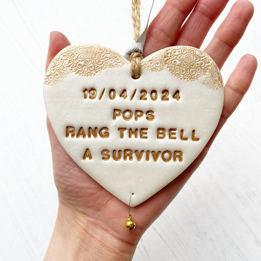 Personalised cancer survivor gift, pearlised white clay hanging heart with a gold lace edge at the top of the heart and a gold bell hanging below, the heart is personalised with 19/04/2024 POPS RANG THE BELL A SURVIVOR