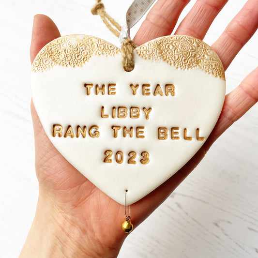 Personalised cancer survivor gift, pearlised white clay hanging heart with a gold lace edge at the top of the heart and a gold bell hanging below, the heart is personalised with THE YEAR LIBBY RANG THE BELL 2023