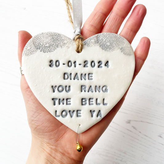 Personalised cancer survivor gift, pearlised white clay hanging heart with a grey lace edge at the top of the heart and a gold bell hanging below, the heart is personalised with 30 - 01-2024 DIANE YOU RANG THE BELL LOVE YA