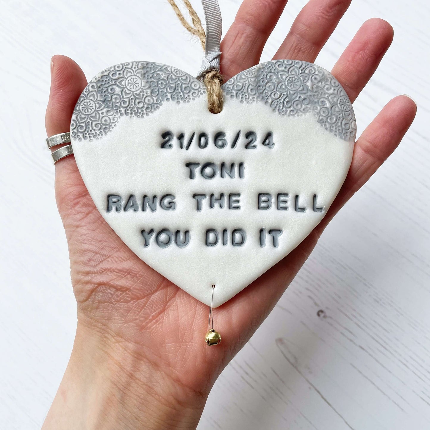 Personalised cancer survivor gift, pearlised white clay hanging heart with a grey lace edge at the top of the heart and a gold bell hanging below, the heart is personalised with 21/06/24 TONI RANG THE BELL YOU DID IT