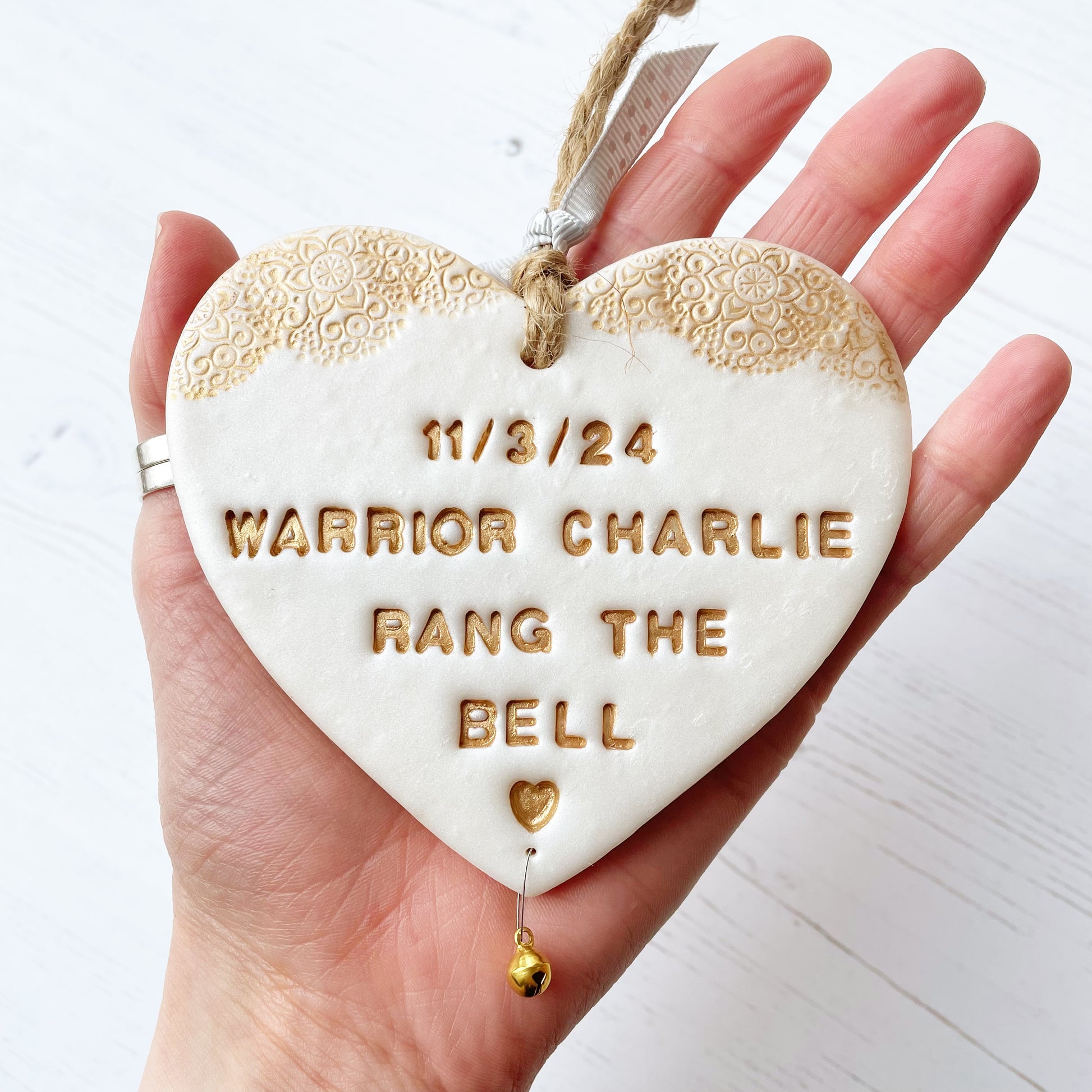 Personalised cancer warrior survivor gift, pearlised white clay hanging heart with a gold lace edge at the top of the heart and a gold bell hanging below, the heart is personalised with 11/3/24 WARRIOR CHARLIE RANG THE BELL 🧡