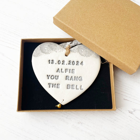 Personalised cancer survivor gift, pearlised white clay hanging heart with a grey lace edge at the top of the heart and a gold bell hanging below, the heart is personalised with 13.02.2024 ALFIE YOU RANG THE BELL. In a luxury Kraft brown gift box