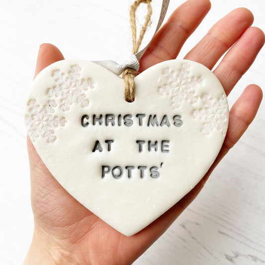 Personalised baby's first Christmas heart ornament, pearlised white clay with CHRISTMAS AT THE POTTS’ painted grey, decorated with 2 iridescent glitter snowflakes on either side of the top of the heart