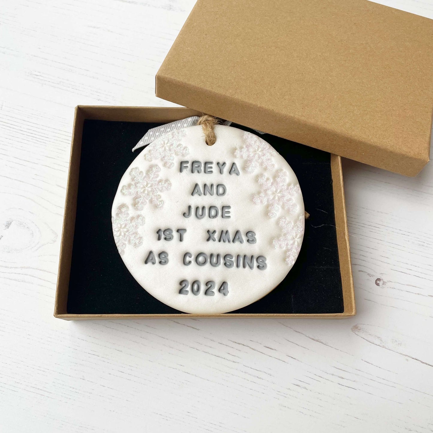 Personalised first Christmas bauble ornament, pearlised white round clay with FREYA SND JUDE 1ST XMAS COUSINS 2024 in grey paint, decorated with 3 iridescent glitter snowflakes on either side of the bauble