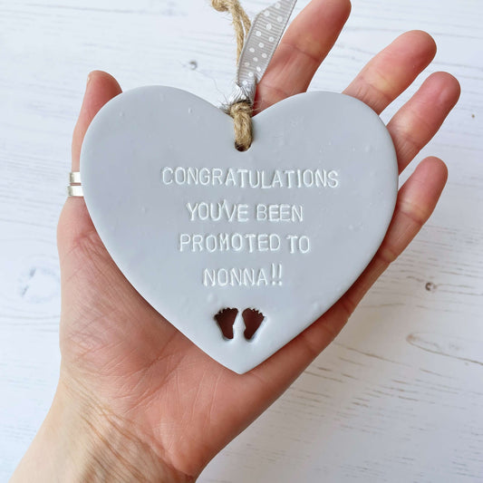 Grey clay hanging heart with baby feet cut out of the bottom and white personalisation, the heart is personalised with CONGRATULATIONS YOU’VE BEEN PROMOTED TO NONNA!!