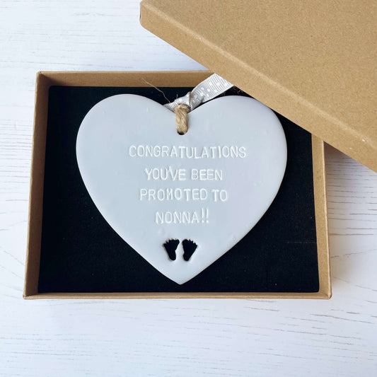 Grey clay hanging heart with baby feet cut out of the bottom and white personalisation, the heart is personalised with CONGRATULATIONS YOU’VE BEEN PROMOTED TO NONNA!!