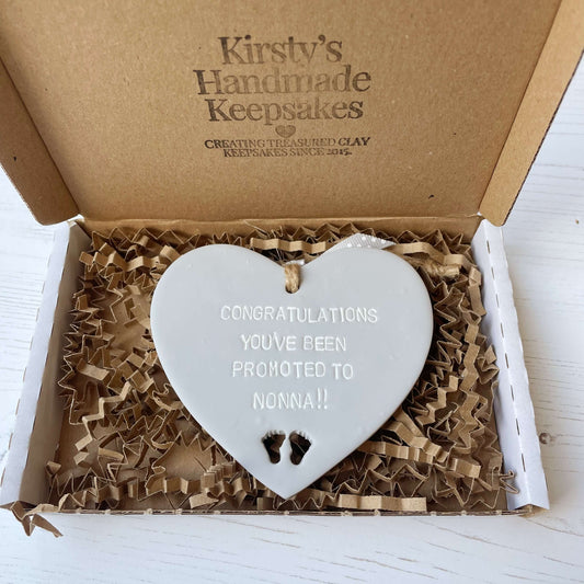 Grey clay hanging heart with baby feet cut out of the bottom and white personalisation, the heart is personalised with CONGRATULATIONS YOU’VE BEEN PROMOTED TO NONNA!!