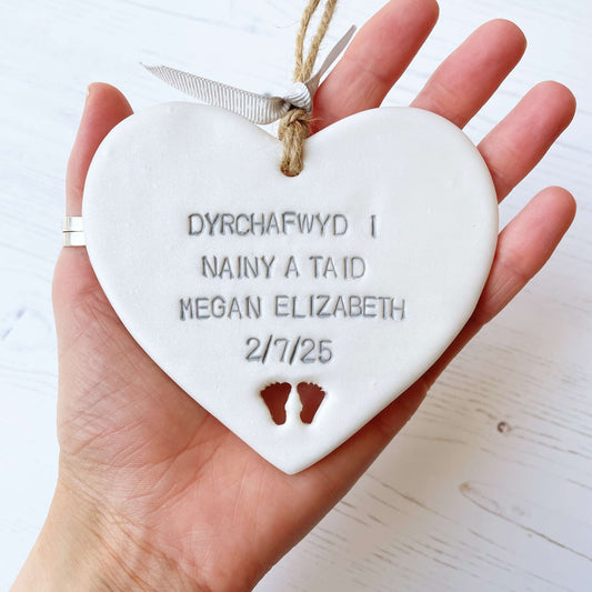 Pearlised white clay hanging heart with baby feet cut out of the bottom and grey personalisation, the heart is personalised with DYRCHAFWYD I NAINY & TAID MEGAN ELIZABETH 2/7/25