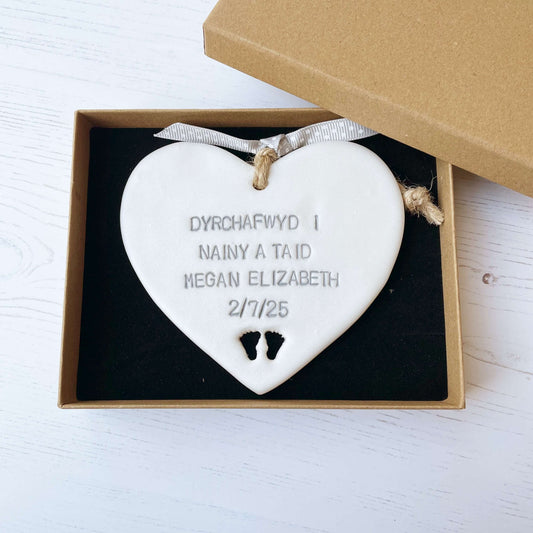 Pearlised white clay hanging heart with baby feet cut out of the bottom and grey personalisation, the heart is personalised with DYRCHAFWYD I NAINY & TAID MEGAN ELIZABETH 2/7/25
