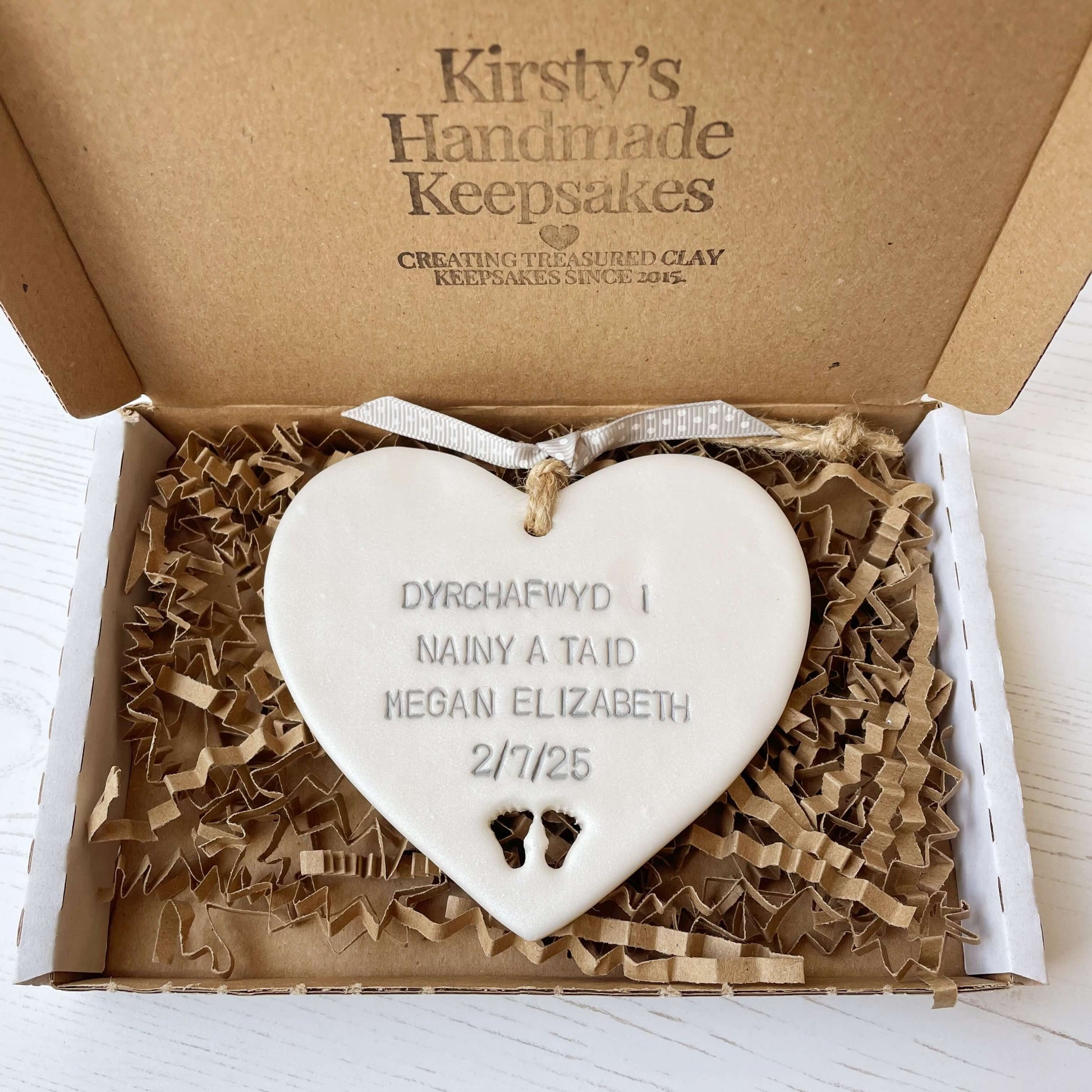 Pearlised white clay hanging heart with baby feet cut out of the bottom and grey personalisation, the heart is personalised with DYRCHAFWYD I NAINY & TAID MEGAN ELIZABETH 2/7/25
